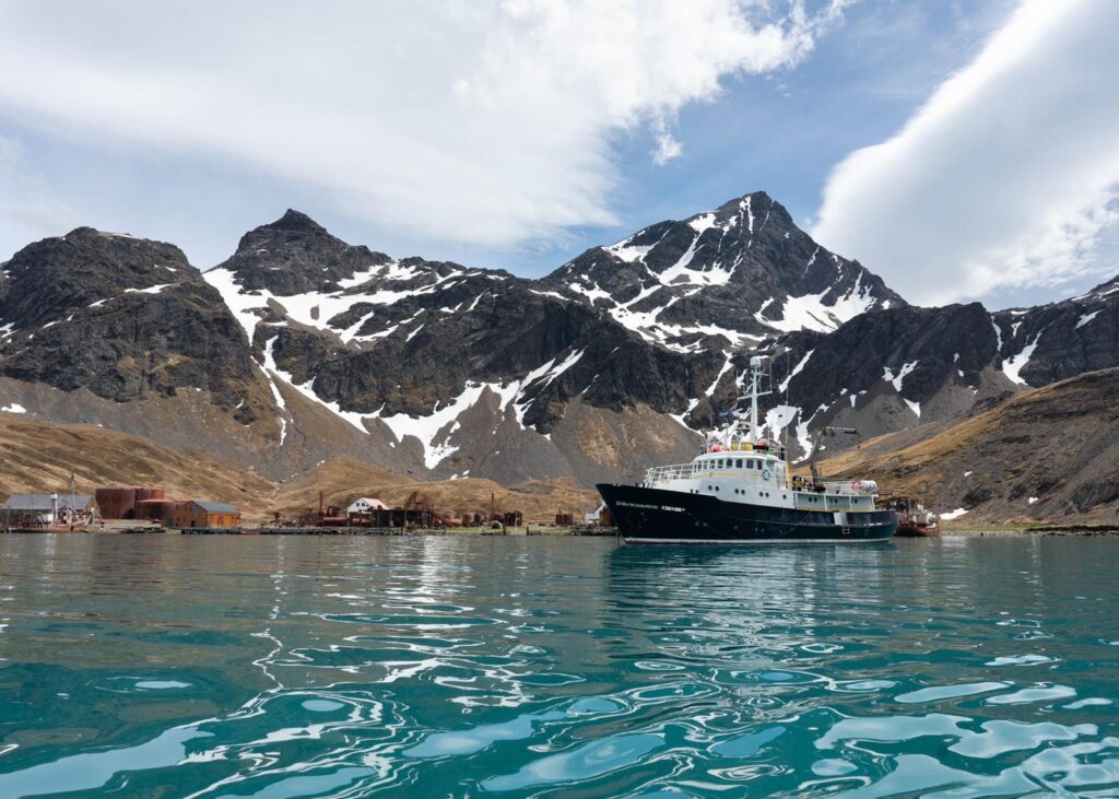 Mountainous landscape with snow patches, boat on water, buildings along shoreline.