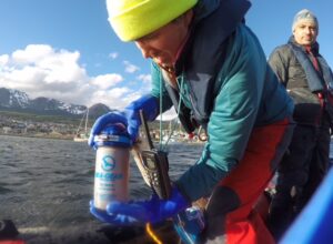 Person in outdoor gear holding SEA-GEAR container and handheld device on a boat in Antarctica, mountains and water in background