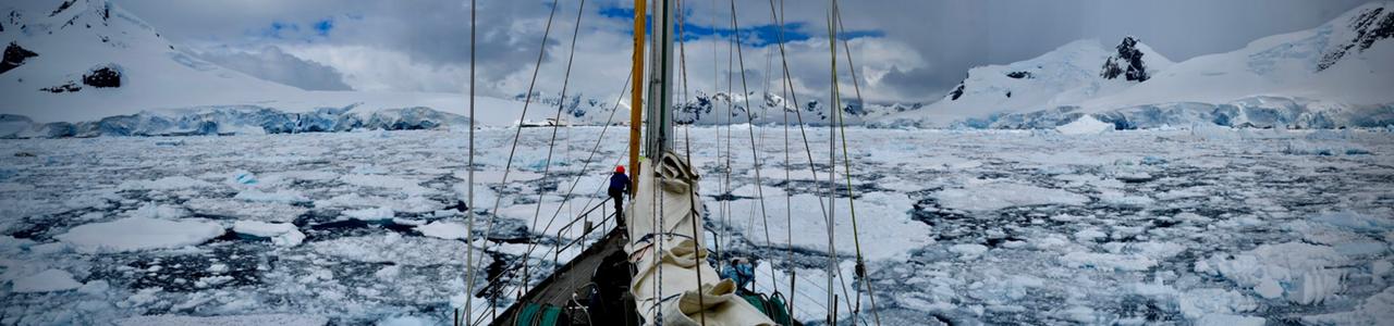 Ship navigating through icy waters in Antarctica with snow-covered mountains in the background, a person standing on the deck.