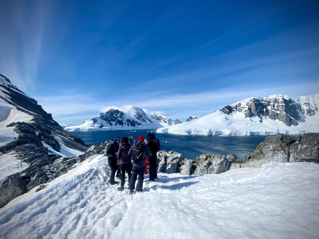 People on a snowy landscape in Antarctica with mountains and water in the background.