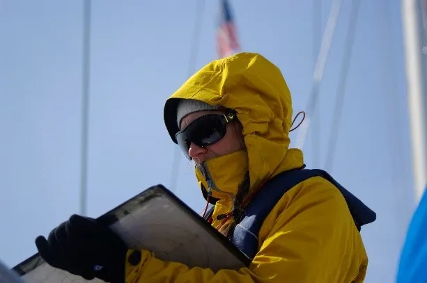 Person in yellow raincoat on boat with clipboard and flag.