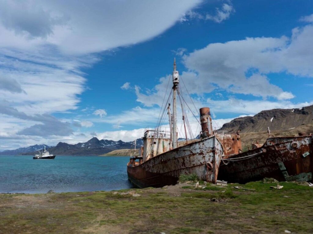 Rusted, abandoned ship on shore with mountains in the background, partly cloudy sky, and distant ship on the water.