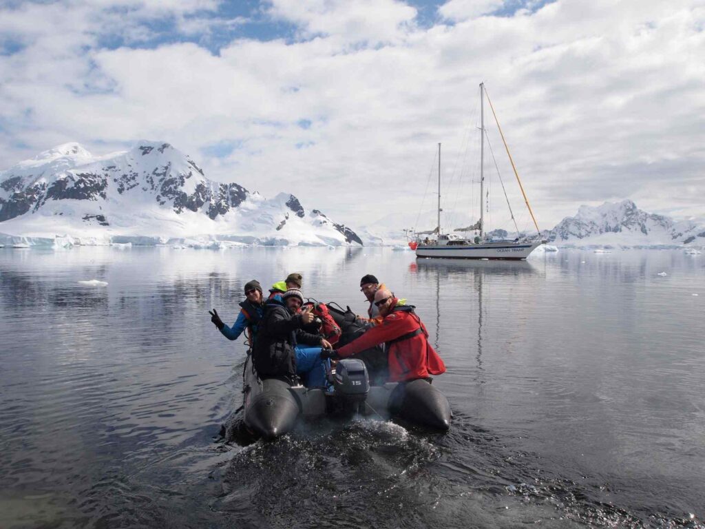 People in inflatable boat observing snow-covered mountains and sailboat in Antarctica.